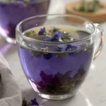 A clear glass cup filled with vibrant butterfly pea flower tea garnished with blue and purple petals.