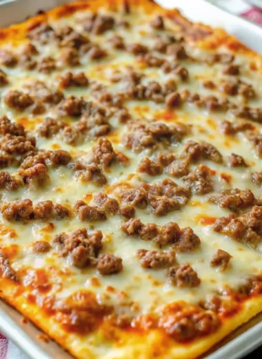Square sausage pizza with melted cheese and ground sausage topping on a baking tray.