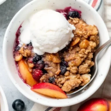 A bowl of Blueberry Peach Crumble dessert with fresh peaches and blueberries, topped with a scoop of creamy vanilla ice cream.